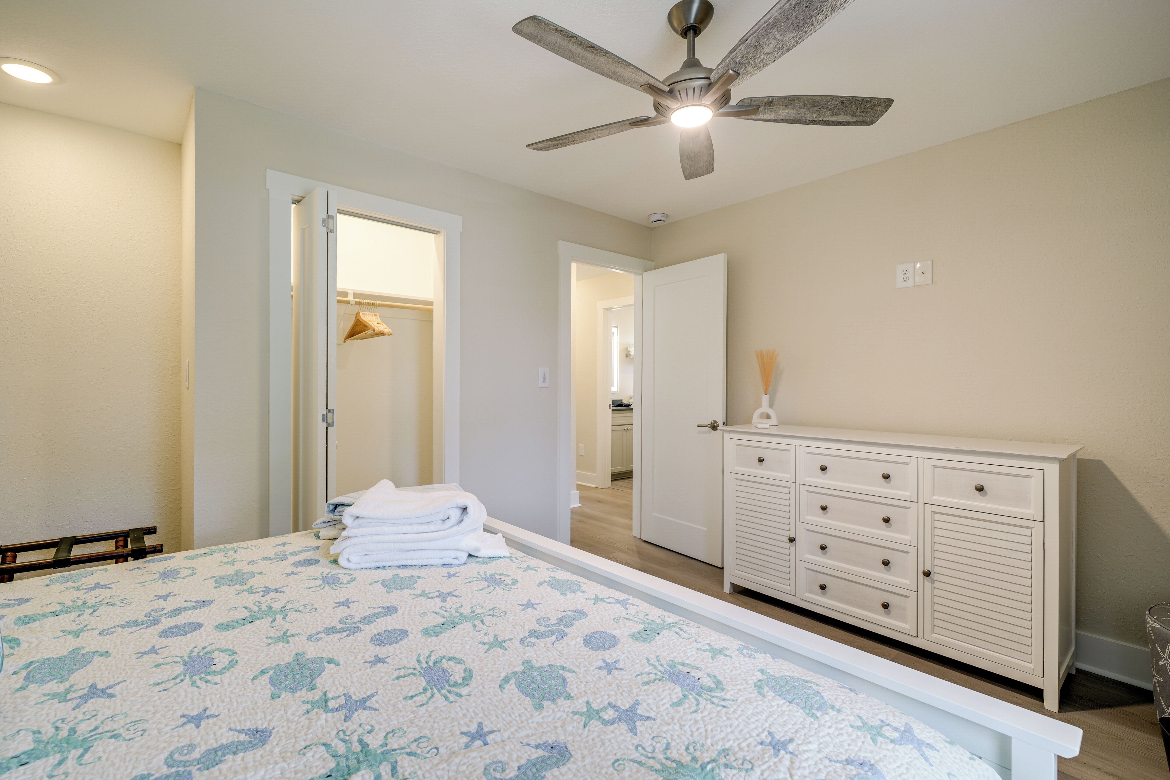 A bedroom with a bed, a dresser, a ceiling fan, and a door leading to another room.