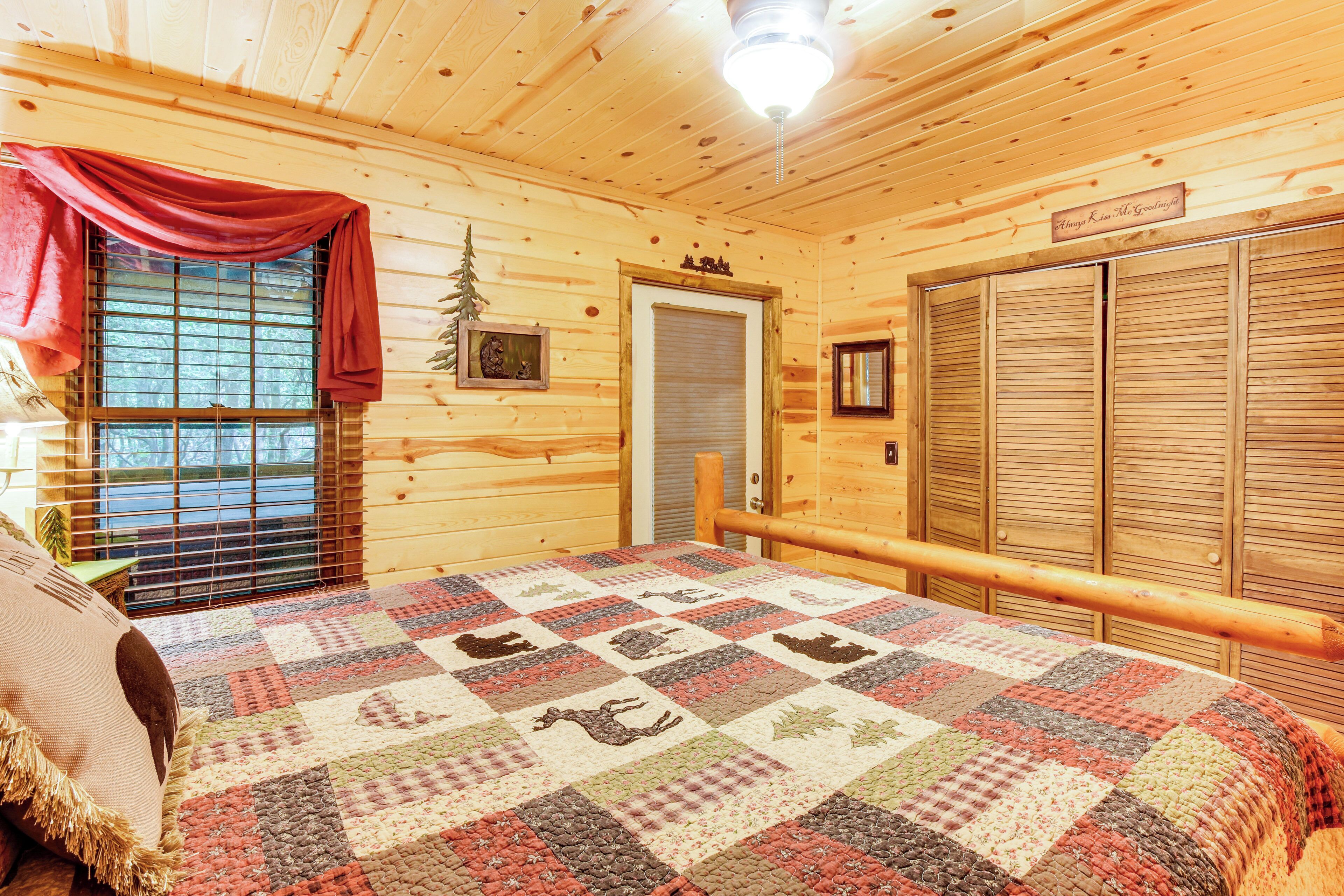 A cozy log cabin room with a quilted bedspread, wooden walls, a window with blinds, and a door with a small tree decoration.