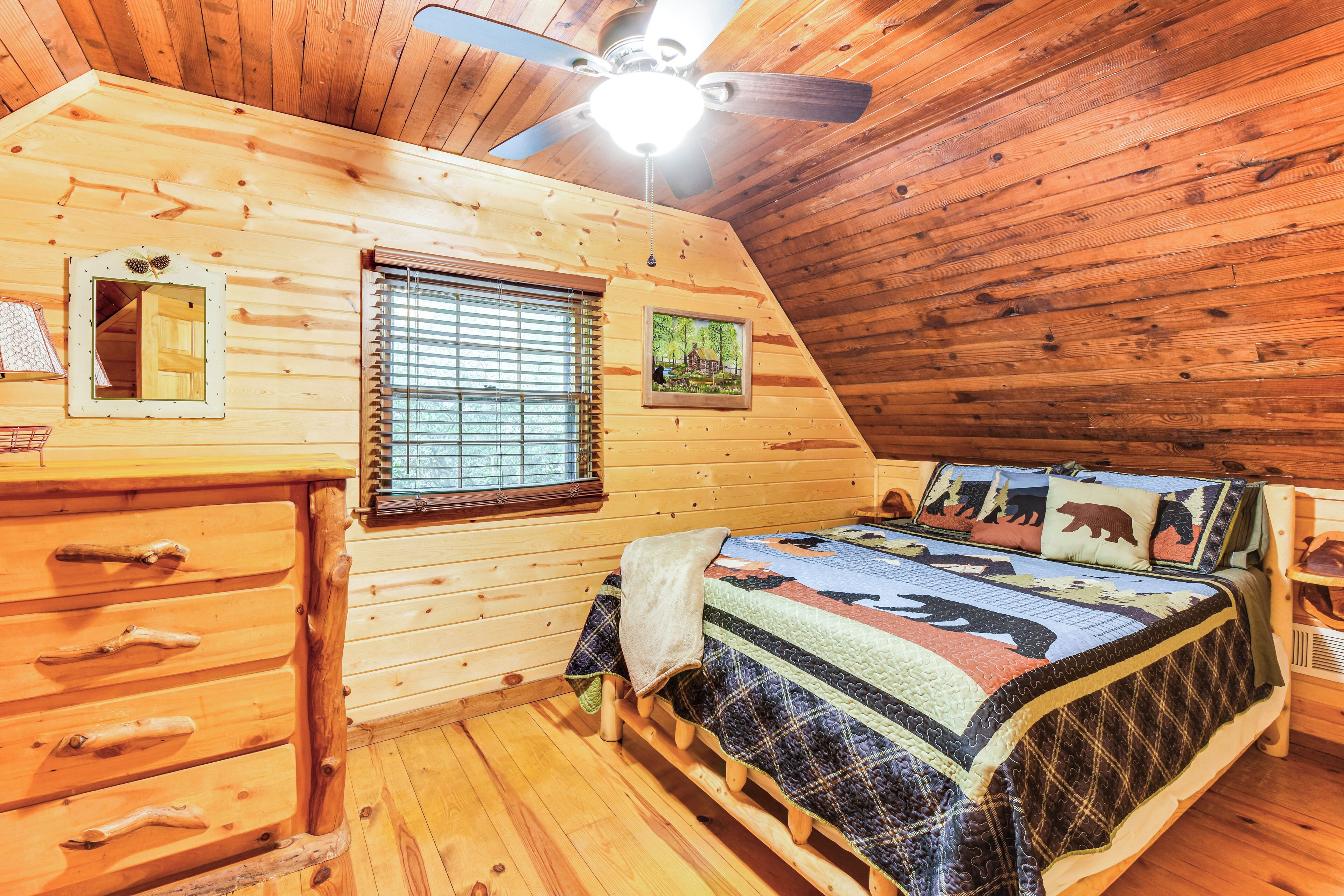 A cozy cabin bedroom with a wooden ceiling, a bed with a quilt, a dresser, a window with blinds, and a ceiling fan.