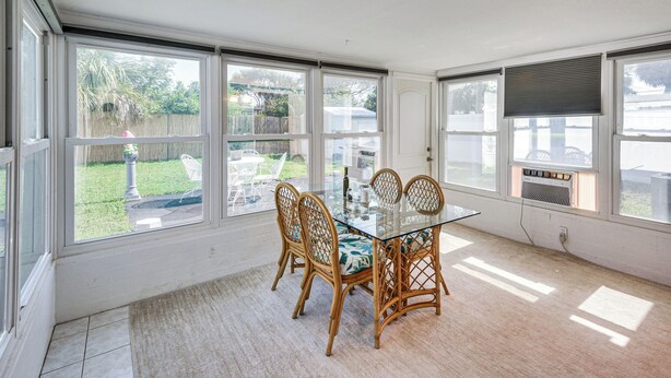 A sunlit room with a glass table and wicker chairs, large windows, and a view of a backyard.