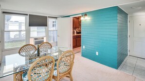 A dining area with a glass table and wicker chairs, a kitchen with wooden cabinets, and a teal-colored wall.