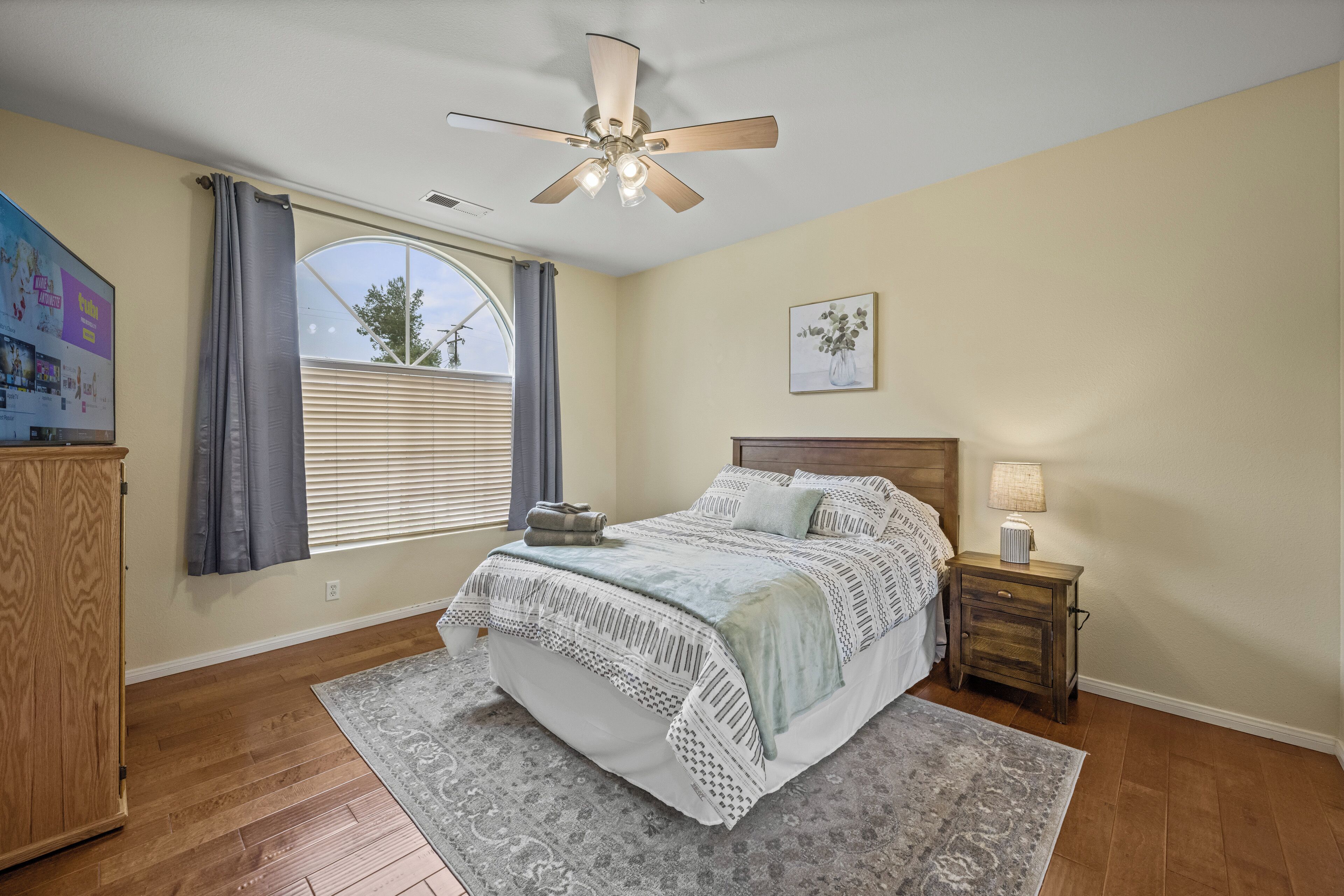 A bedroom with a bed, a nightstand, a ceiling fan, a window with curtains, and a wooden wardrobe.