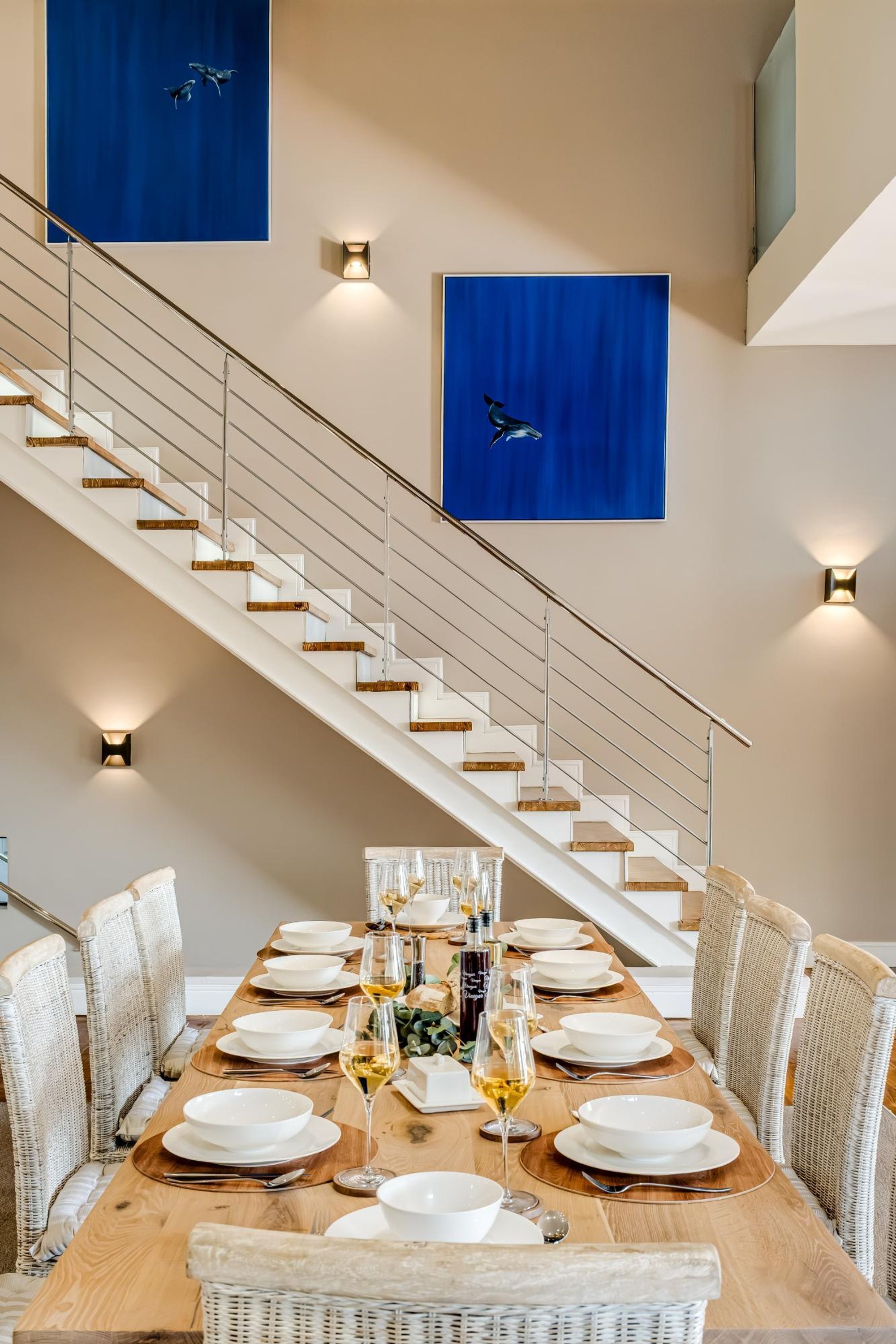 A dining room with a long table set for a meal, a staircase, and two blue artwork pieces on the walls.