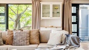 A modern living room with a sofa, a cabinet with glass doors, and a view of a tree outside. - 2bed Apt 5min Walk From Camps Bay Central Escape (Cape Town)
