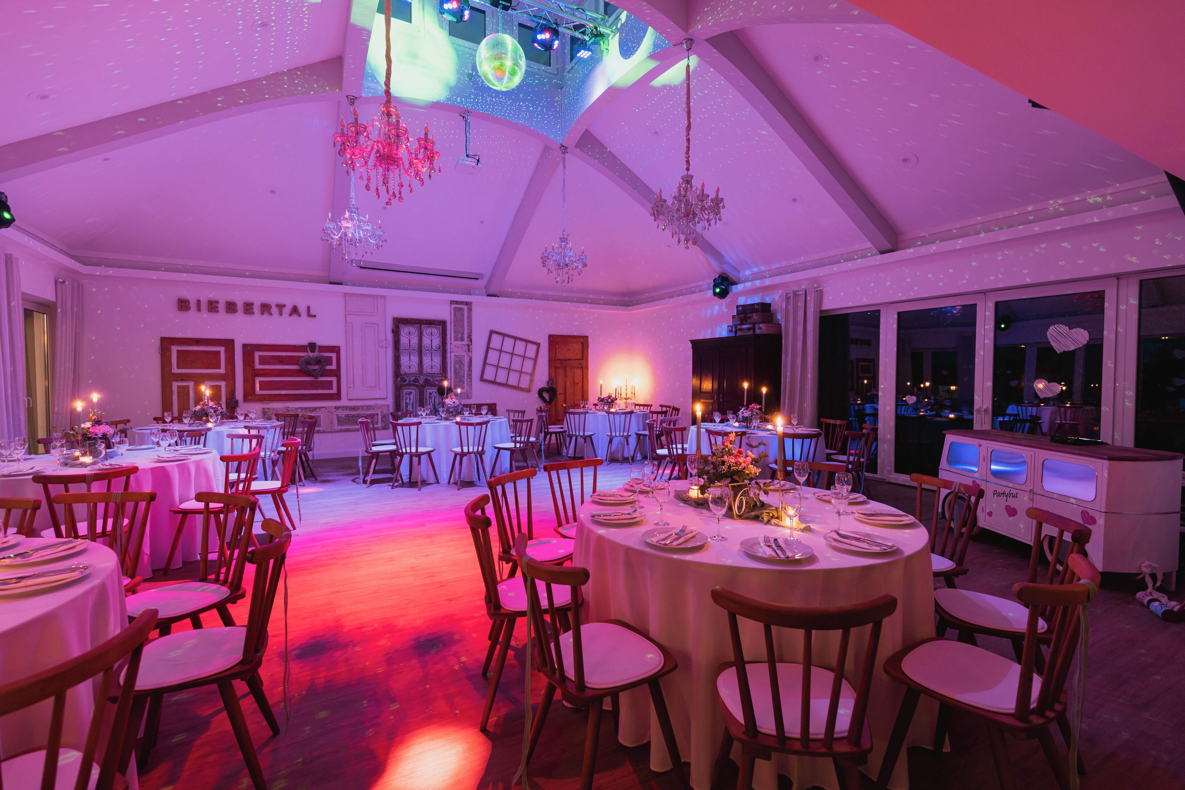 A spacious banquet hall with round tables, wooden chairs, and a high ceiling with chandeliers.
