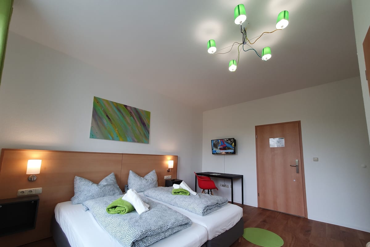 Ein Doppelbett mit gemusterten Betttextilien, ein Nachttisch mit Lampe, ein an der Wand befestigter Fernseher und eine Holztür.
