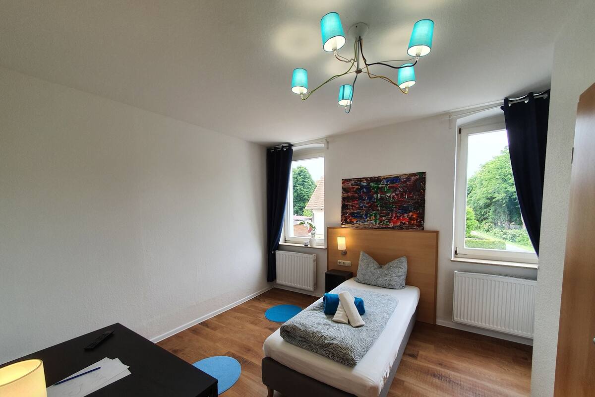 Ein Schlafzimmer mit einem Bett, einem Schreibtisch, einem Stuhl, einer Lampe und einer Deckenleuchte.