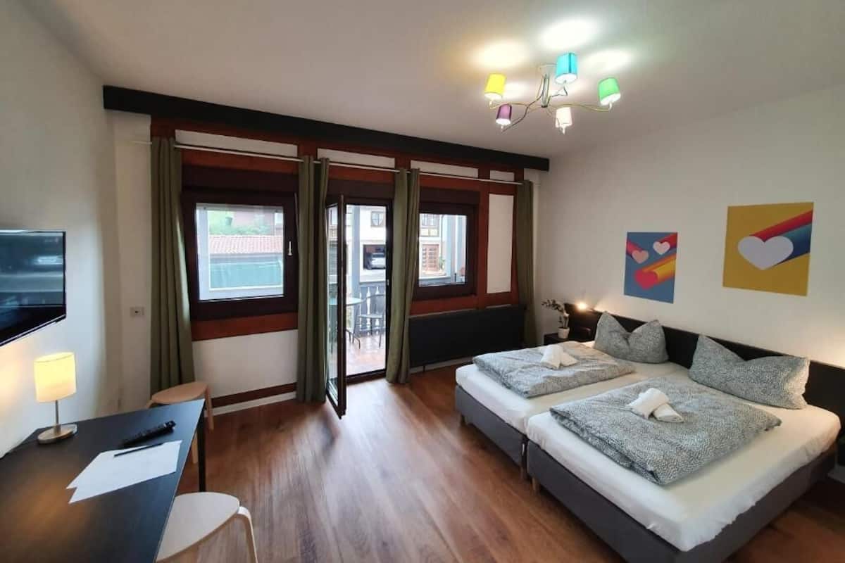 Ein Hotelzimmer mit zwei Betten, einem Schreibtisch, einem Fernseher und einem Balkon mit Vorhängen.