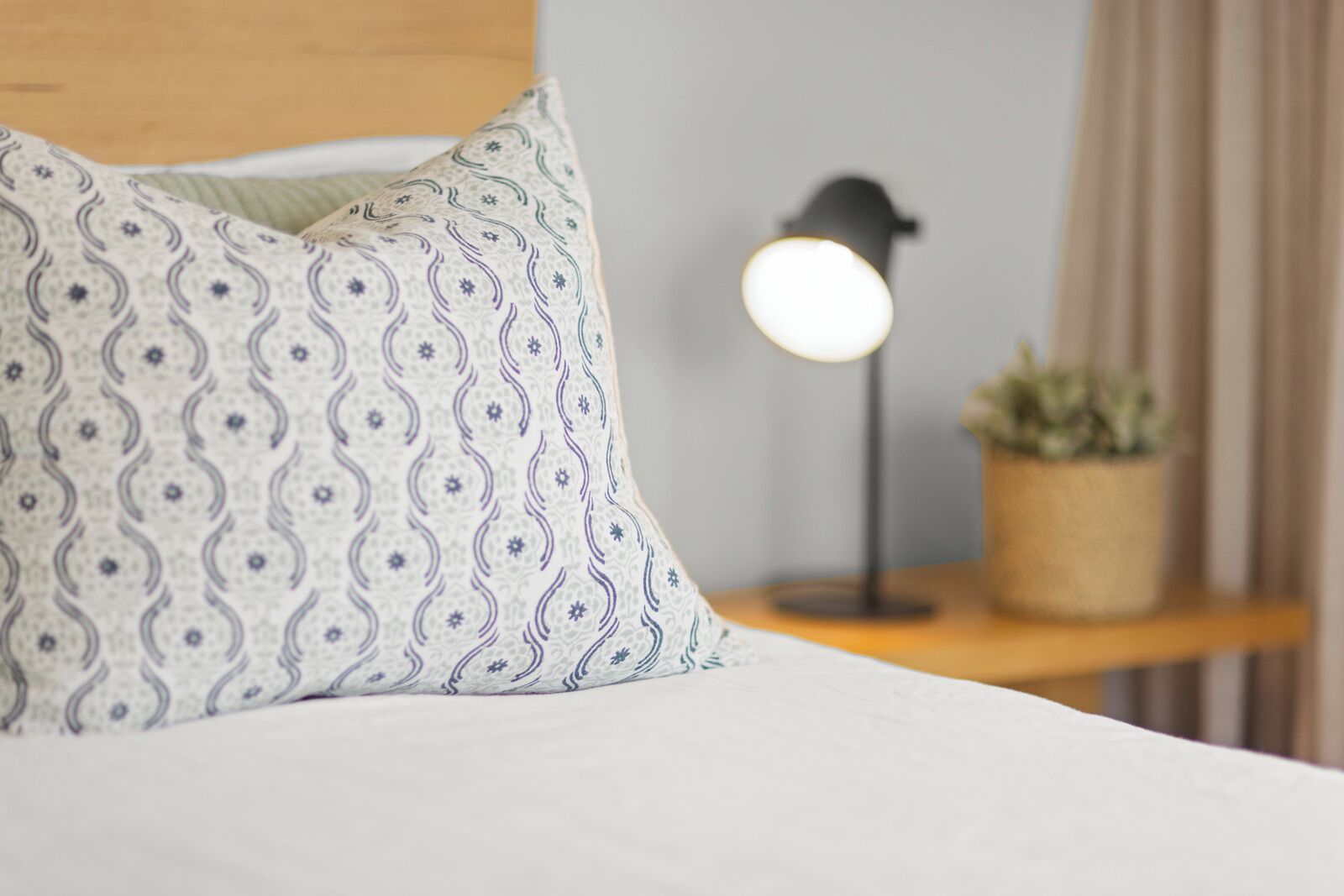 A neatly made bed with a patterned pillow, a bedside lamp, and a small plant on a wooden nightstand.