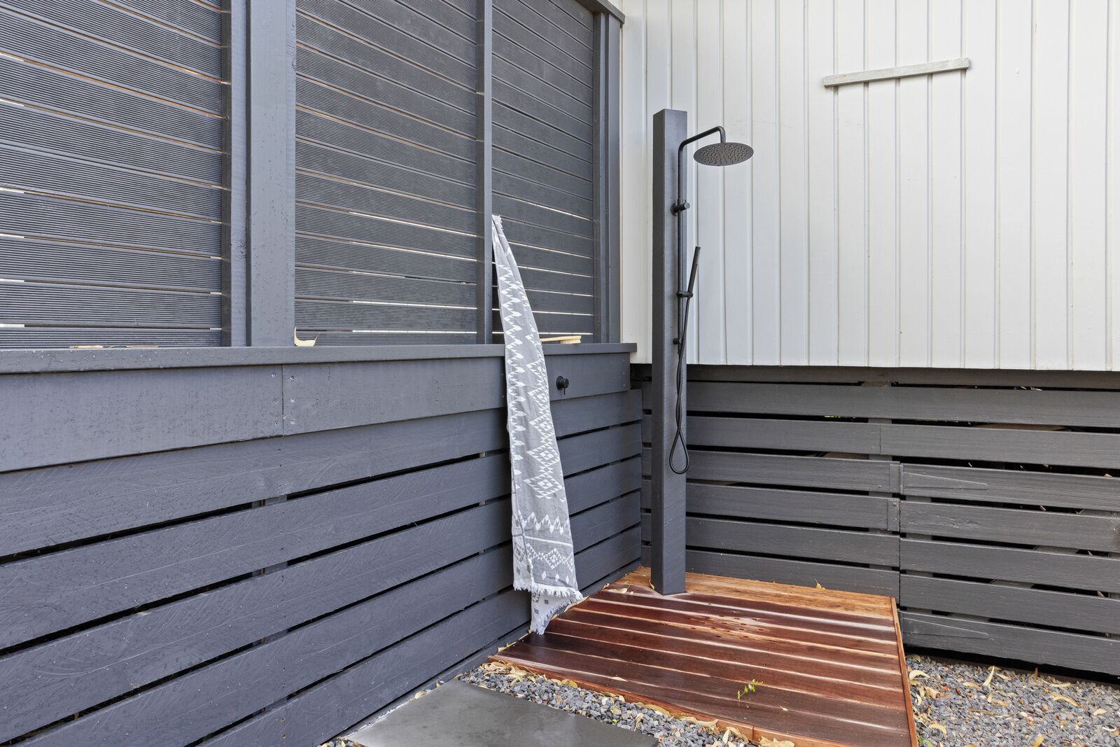 A modern outdoor shower area with a metal hose and a showerhead, adjacent to a building with dark wooden siding and a window with horizontal blinds.