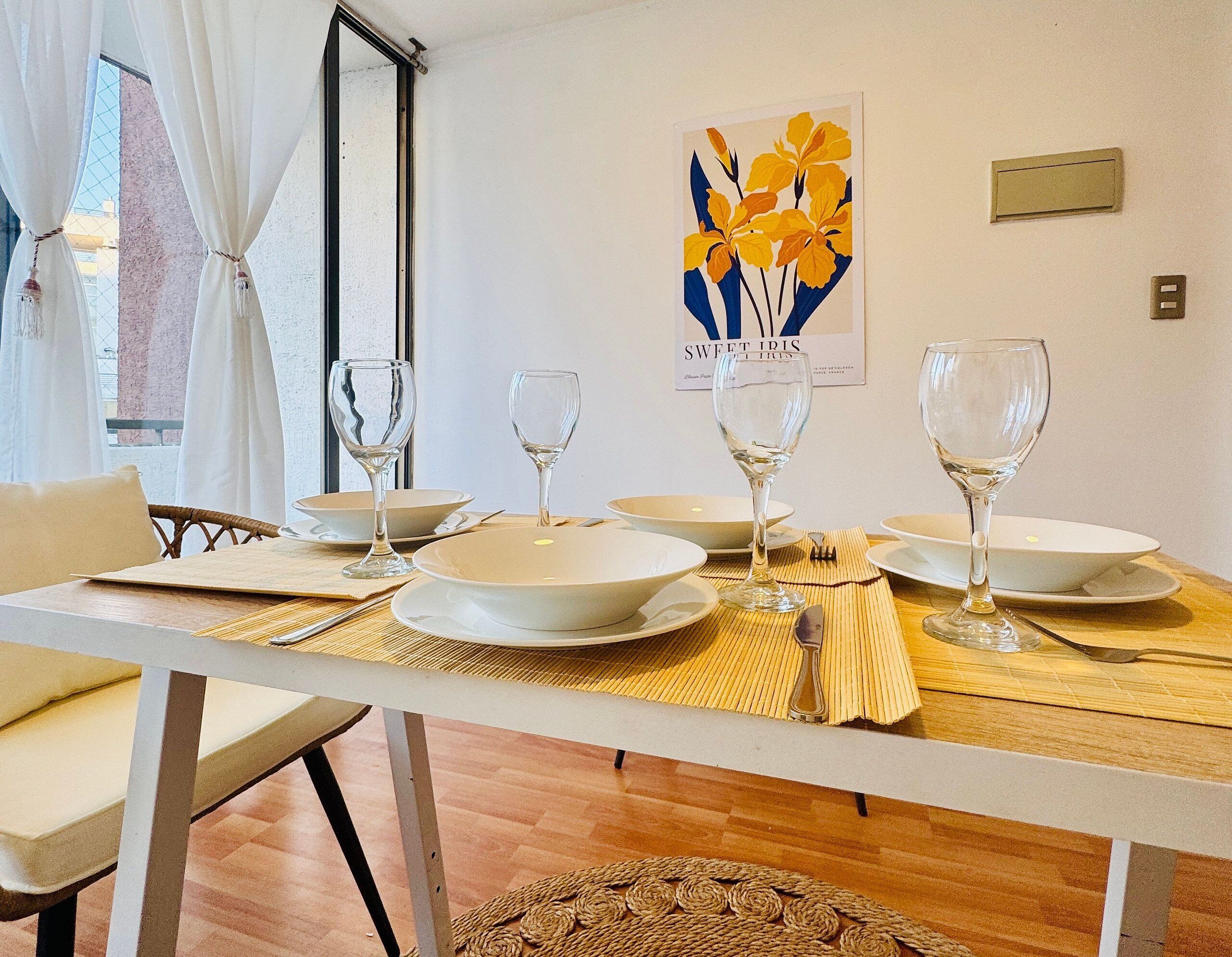 Een eettafel gedekt met witte borden en wijnglazen, een geweven placemat en een rieten stoel.