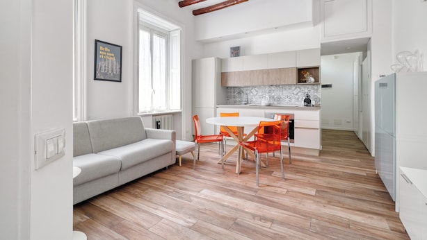 A modern kitchen with white cabinets, a wooden floor, and a dining area with a white table and orange chairs. - Lovely flat near Naviglio Grande (Milan)