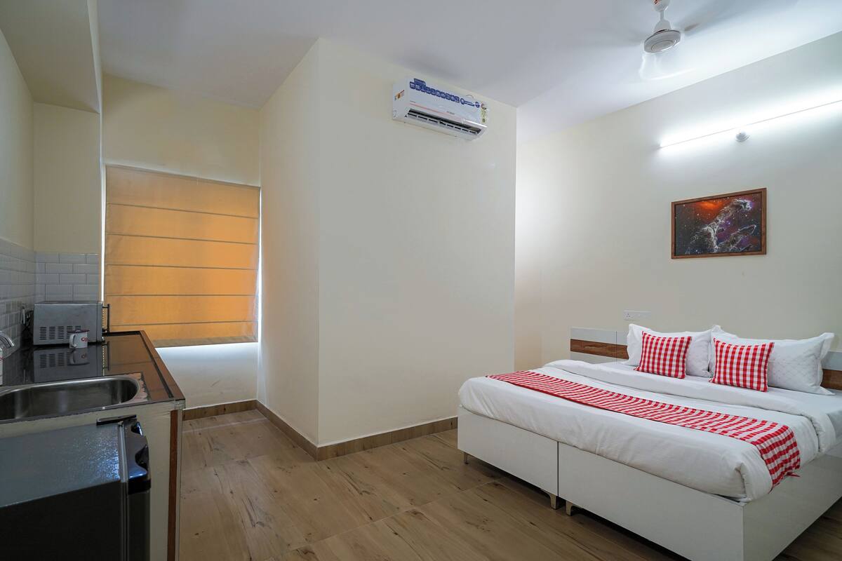 A hotel room with a bed, a small kitchenette, a ceiling fan, and a window with blinds.