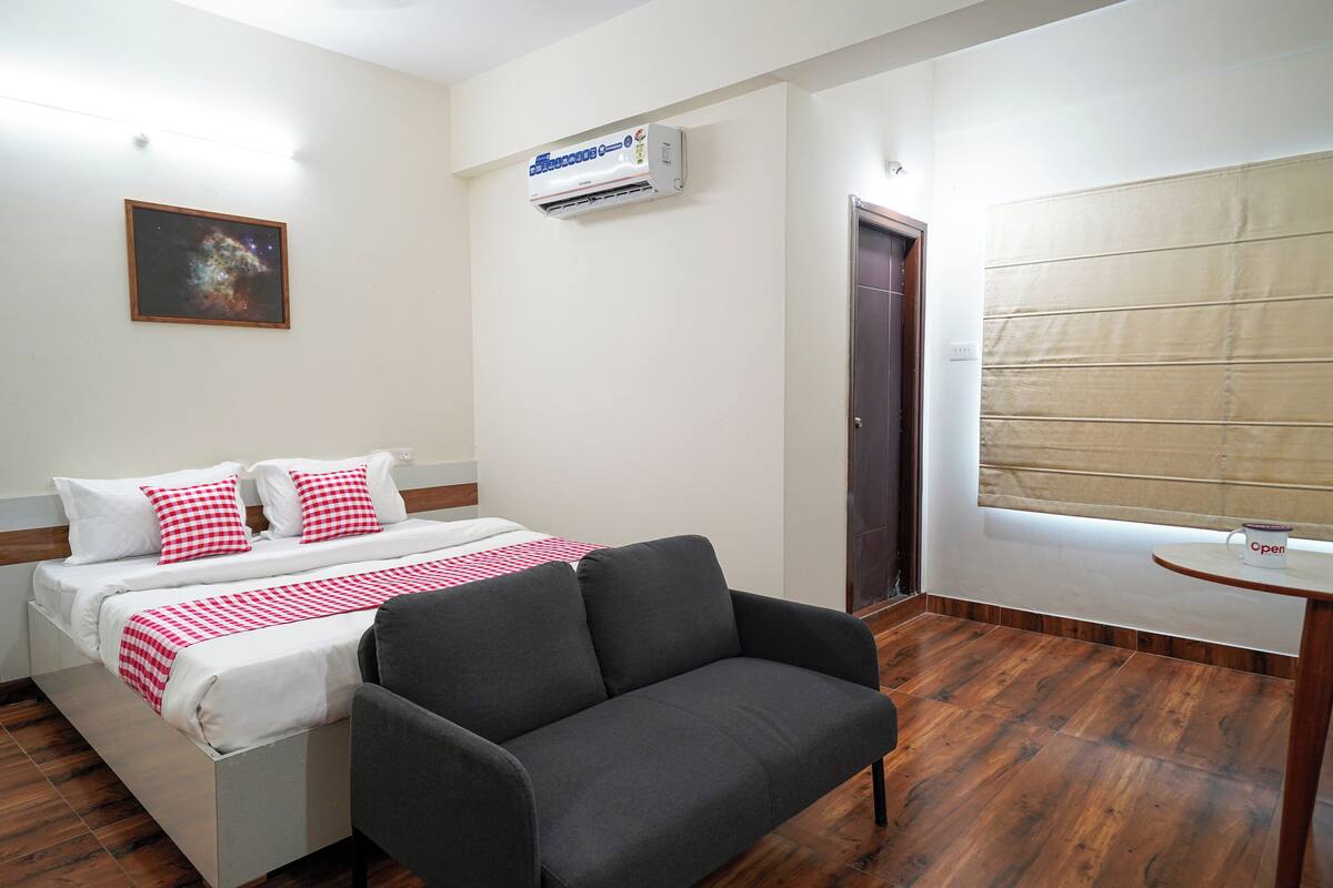 A hotel room with a bed, a gray armchair, a window with blinds, an air conditioning unit, and a door.