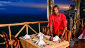 A dining area with a table set for two, wooden chairs, and a view of the ocean at dusk. - Mara Sun Lodge (Maasai Mara)