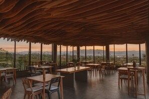 Un área de comedor al aire libre con mesas y sillas de madera, y amplios ventanales con vistas a las montañas.