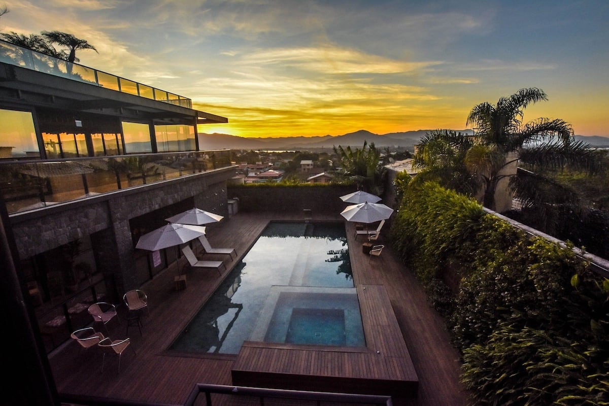 A rooftop pool with lounge chairs and umbrellas, surrounded by greenery and overlooking a cityscape at sunset.