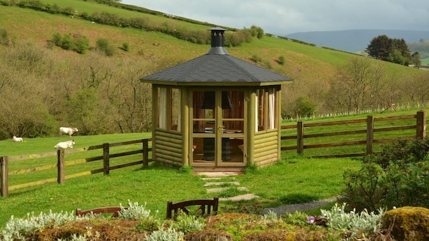 A small wooden cabin with a glass door, surrounded by a wooden fence and a grassy field with cows grazing. - Tucked Away Log Cabin on Welsh Hill Farm (Sennybridge)