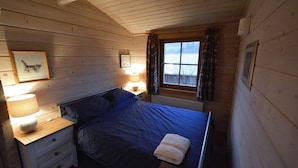 A bedroom with a wooden ceiling, a bed with a blue mattress, a nightstand with a lamp, and a window with curtains. - Tucked Away Log Cabin on Welsh Hill Farm (Sennybridge)