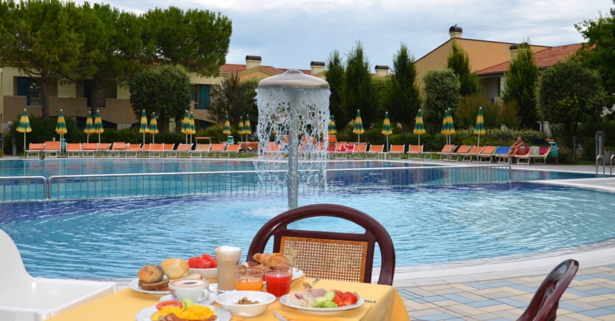 A pool with a fountain, surrounded by lounge chairs and umbrellas, with breakfast on a table.
