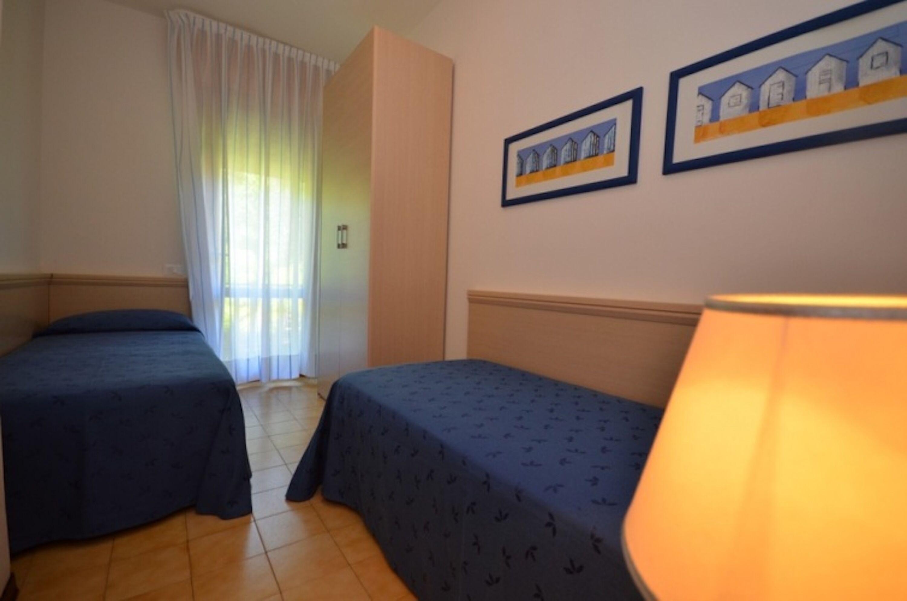 Quarto de hotel com duas camas individuais, uma janela com cortinas leves e duas quadros na parede.