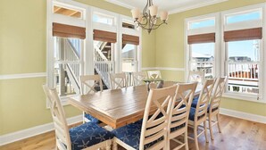 A dining room with a wooden table and chairs, large windows with blinds, and a chandelier. - The Penalty Box- 5 bed, 5+ bath with putting green, corn hole, & hammock swing. (Ocean Isle Beach)