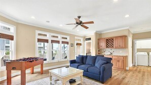 A living room with a foosball table, a blue sofa, and a ceiling fan. - The Penalty Box- 5 bed, 5+ bath with putting green, corn hole, & hammock swing. (Ocean Isle Beach)