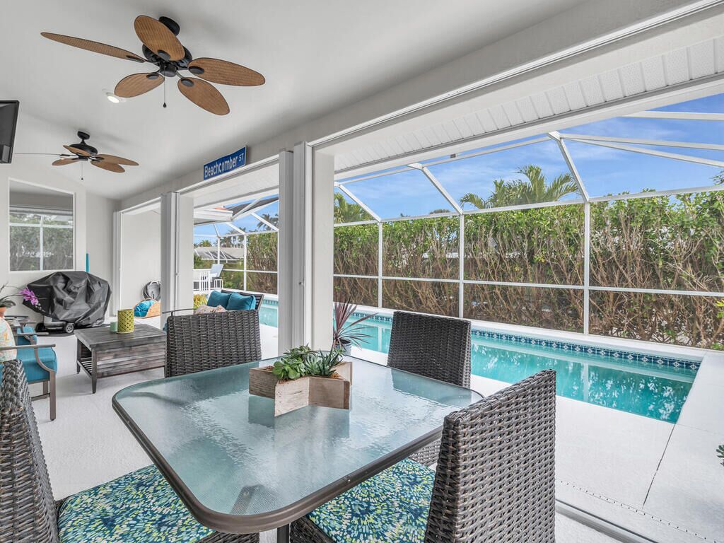 A sunlit room with a glass table, wicker chairs, a ceiling fan, and a view of a swimming pool.