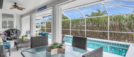 A sunlit room with a glass table, wicker chairs, a ceiling fan, and a view of a swimming pool.