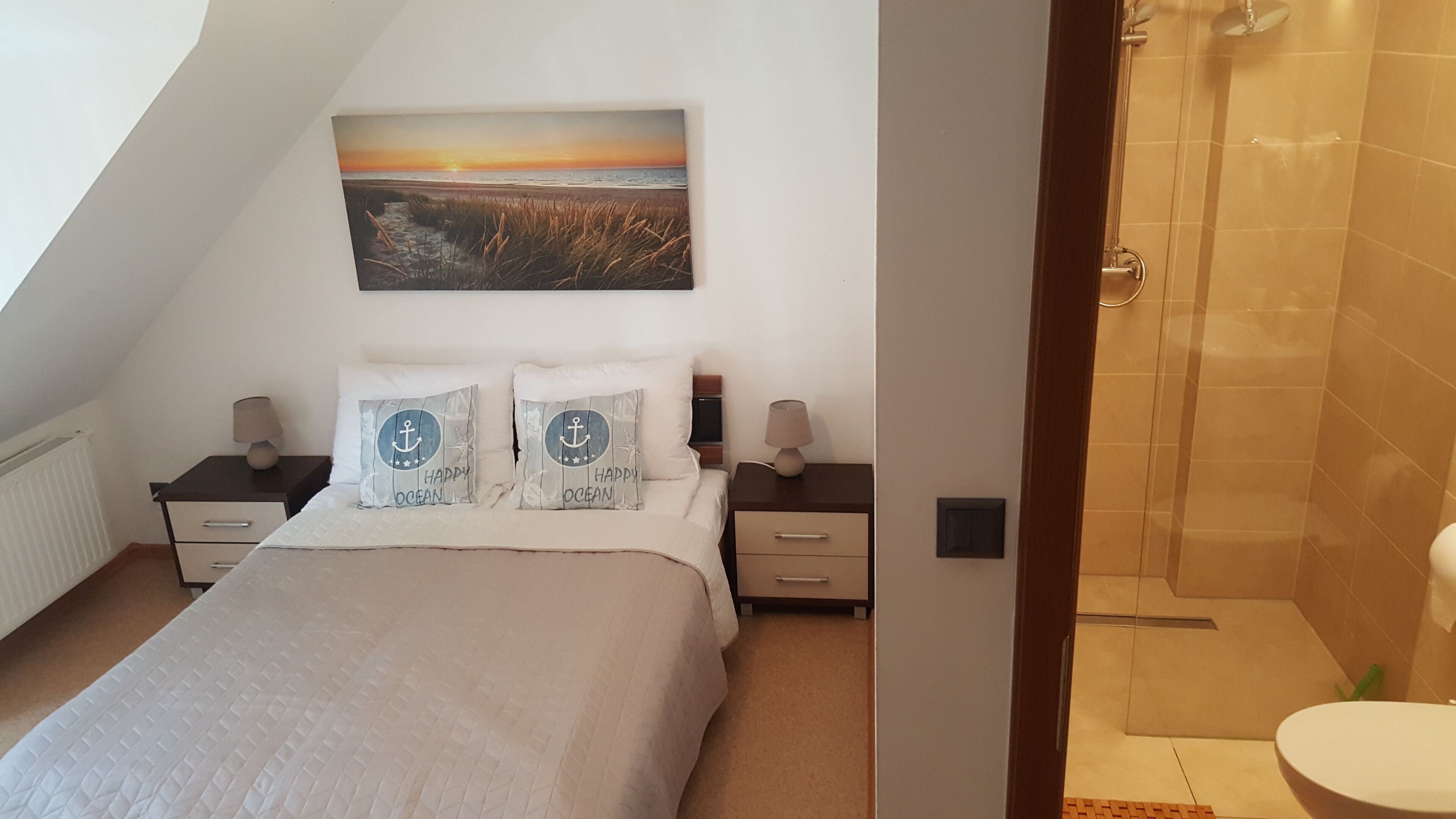A bedroom with a bed, two bedside tables, a nightstand, a painting, and a shower area.
