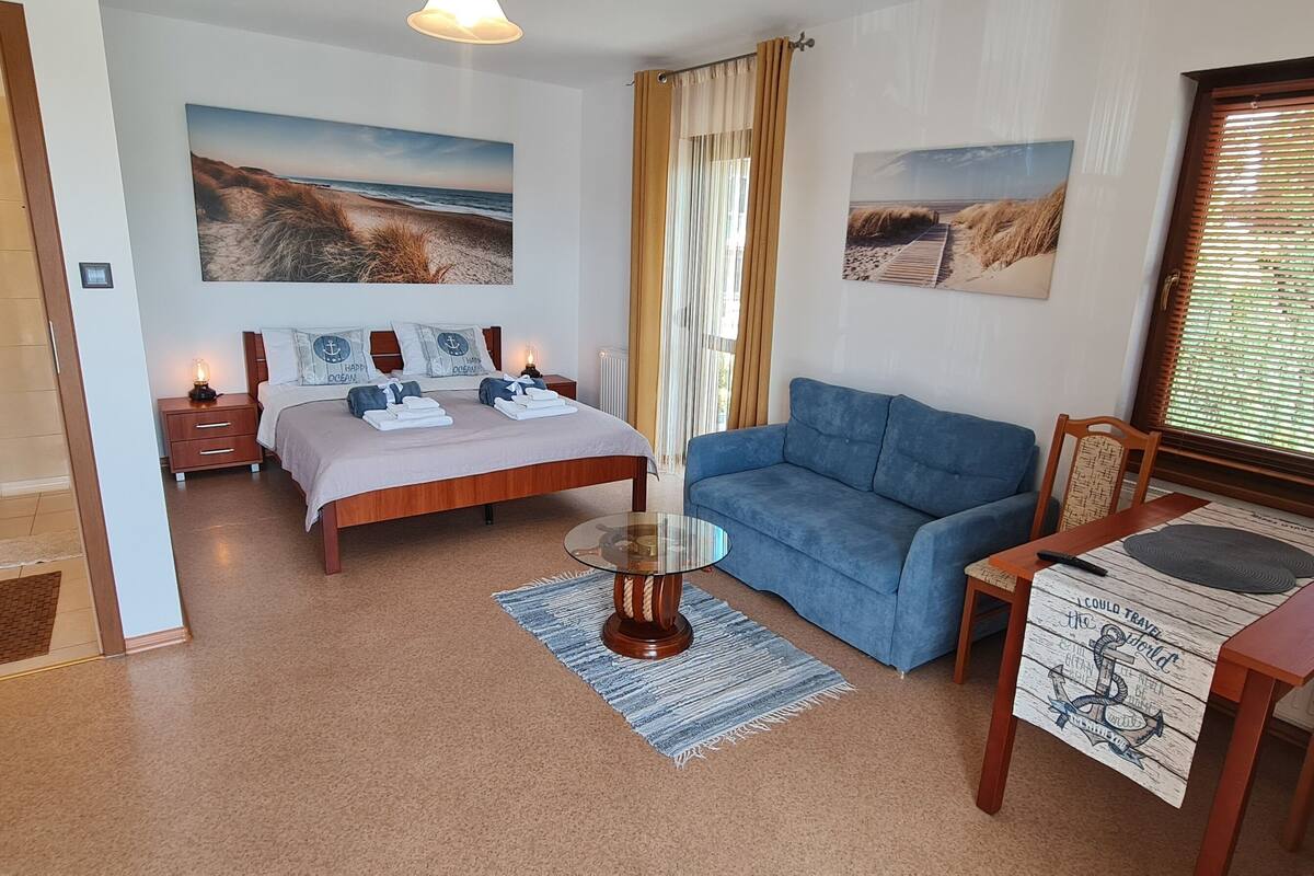 Ein Schlafzimmer mit einem Bett, einem blauen Sofa, einem Glastisch, einem Holznachttisch und einem Fenster mit Jalousien.