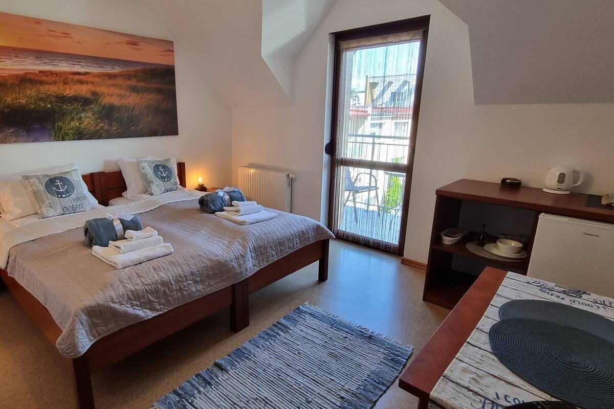 Ein Schlafzimmer mit einem großen Bett, Nachttischen mit Lampen, Blick ins Freie durch ein Fenster und einer kleinen Küchenzeile.