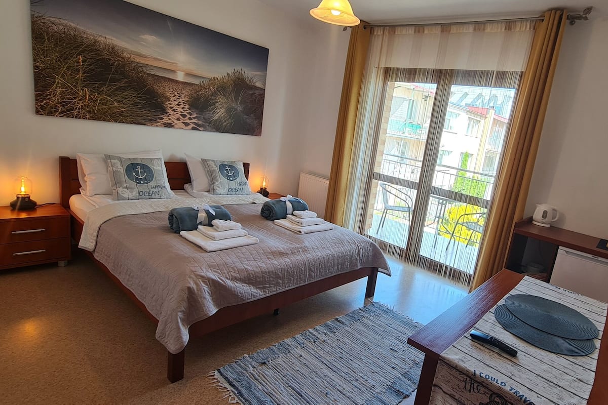 Ein Schlafzimmer mit Bett, Nachttischen, Gemälde, einem Fenster mit Vorhängen und Blick auf Gebäude.