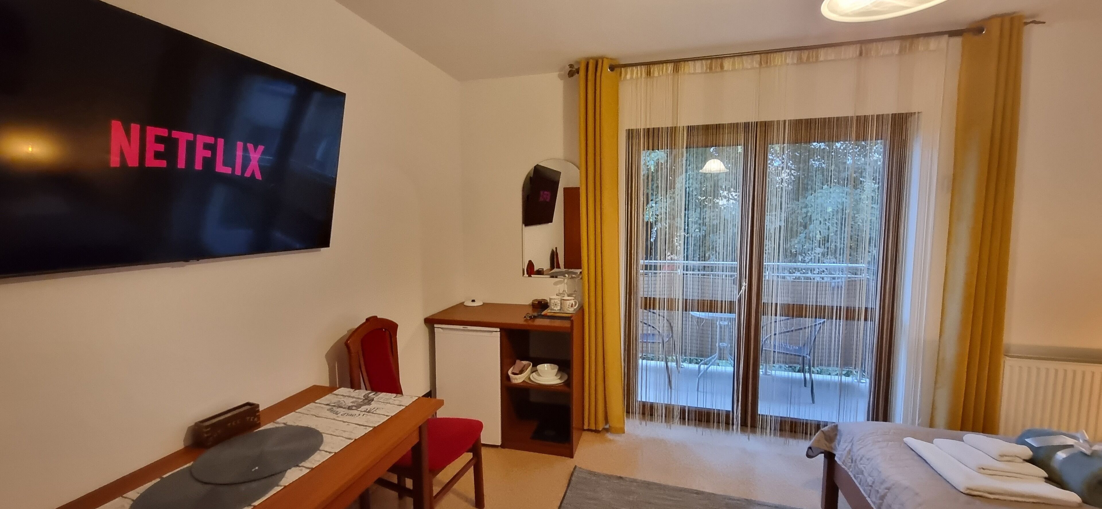 Una habitación con televisión mostrando el logotipo de Netflix, una kitchenette pequeña, una zona de comedor con mesa y sillas, y un ventanal corredero que da a un balcón exterior.