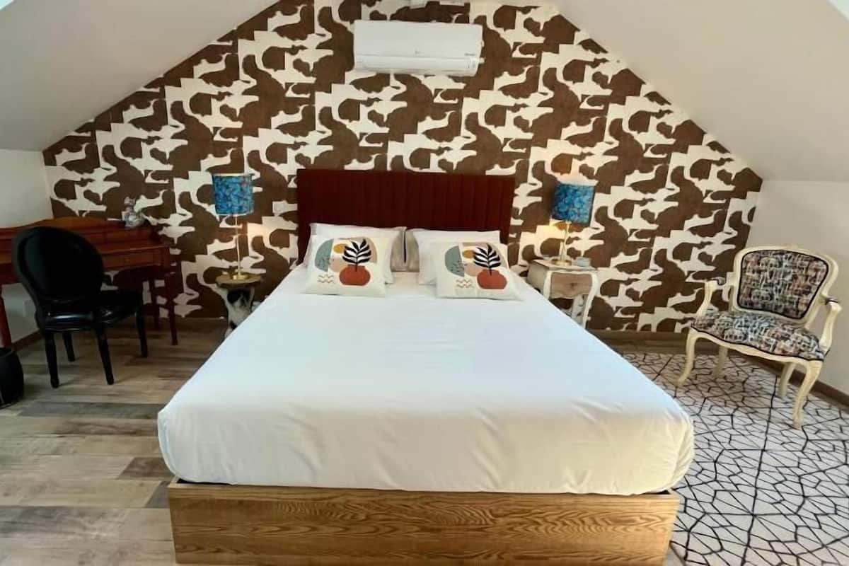 A bedroom with a sloped ceiling, a bed with white bedding, a wooden headboard, a desk with a chair, a small table with a lamp, and a patterned wall.