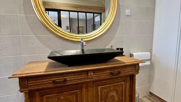 A bathroom with a wooden vanity, a black vessel sink, and a round mirror with a gold frame. - Cave Auger La Maison d'Hotes (Restigne)