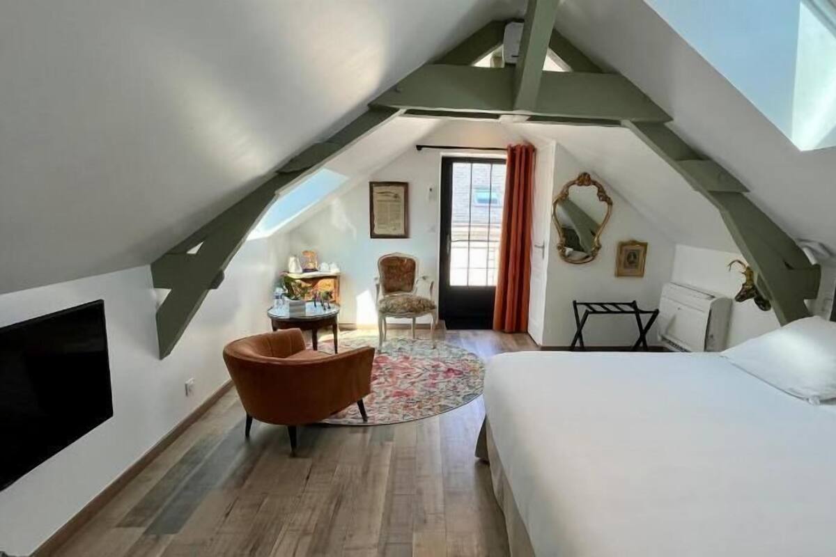 A modern attic bedroom with a large bed, a seating area with a chair, and a TV.