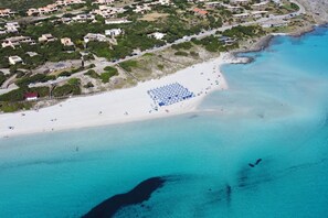 Vue aérienne d’une zone côtière avec une plage, des bâtiments et une eau d’un bleu limpide.