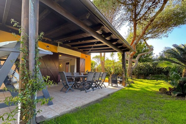 Un patio al aire libre con una mesa y sillas, rodeado de follaje y un edificio con un área cubierta.