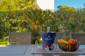 A table with a bottle of Berlucci wine, a bowl of fruit, and glasses, set outdoors with chairs and greenery in the background.