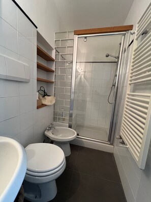Et badeværelse med toilet, bidet og brusebad. Der er hylder med toiletartikler og en håndklæderække.