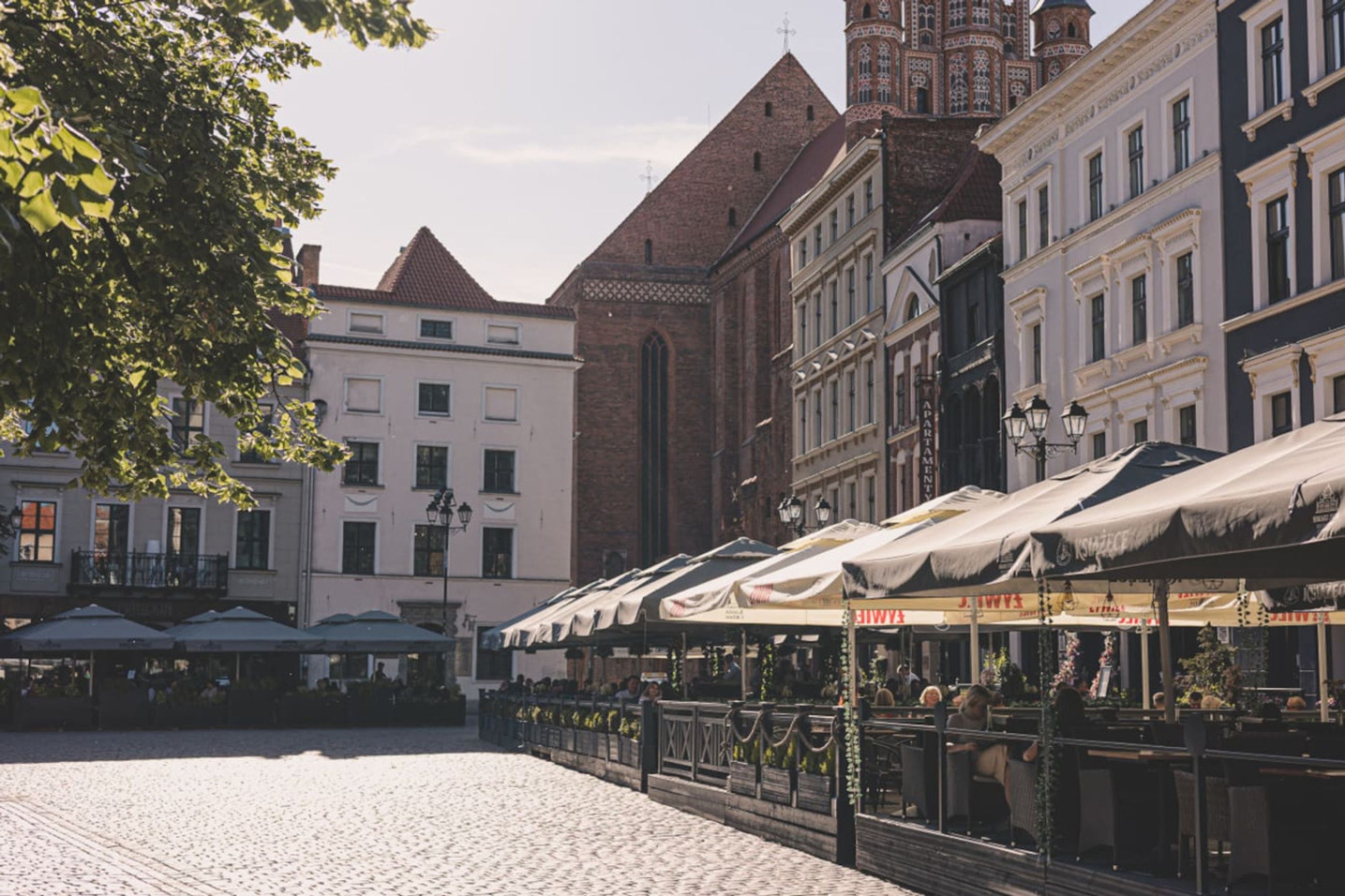 A cobblestone square with outdoor seating under umbrellas, historic buildings, and a church with a tall spire.