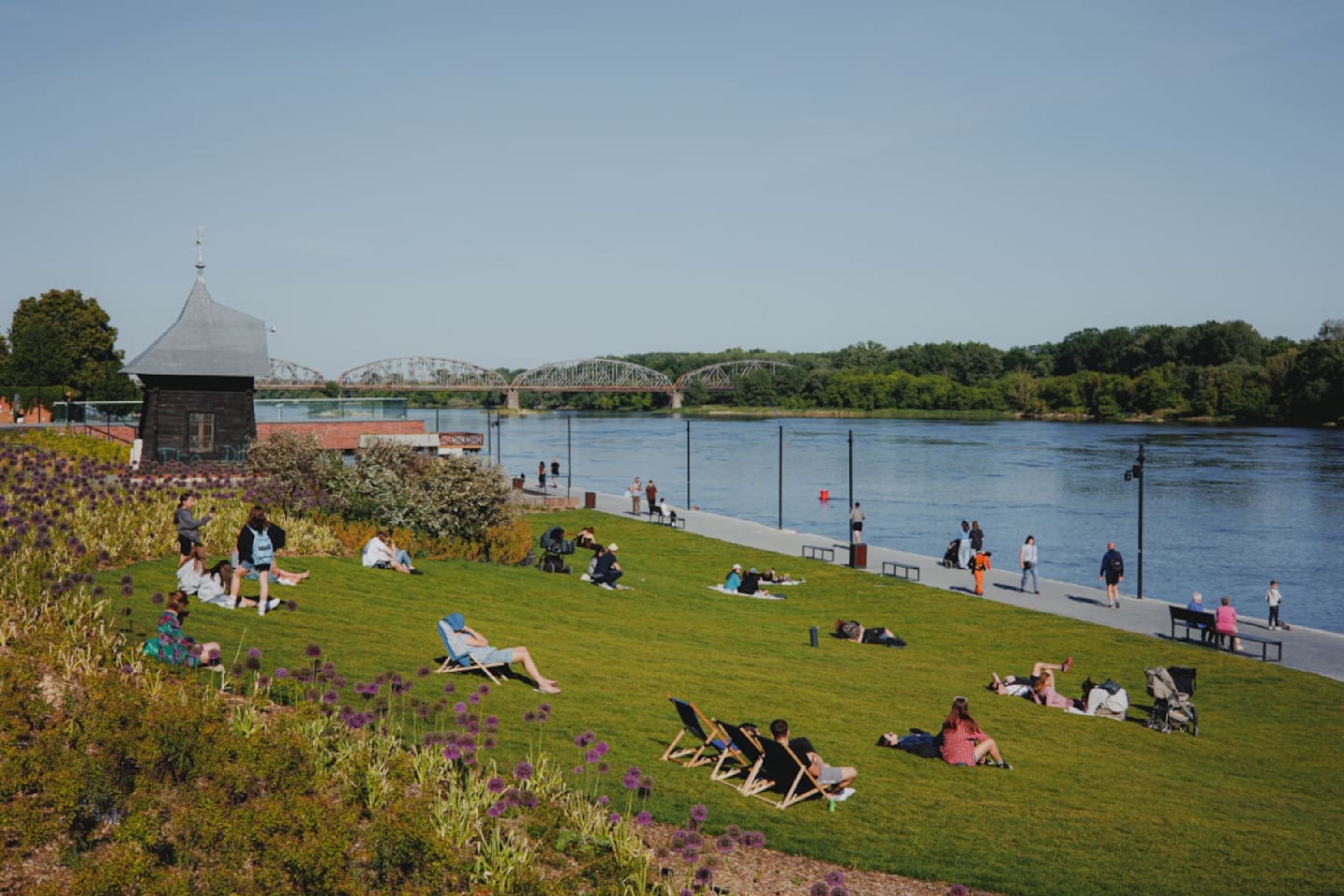 A riverside park with grassy areas, benches, and a small building.