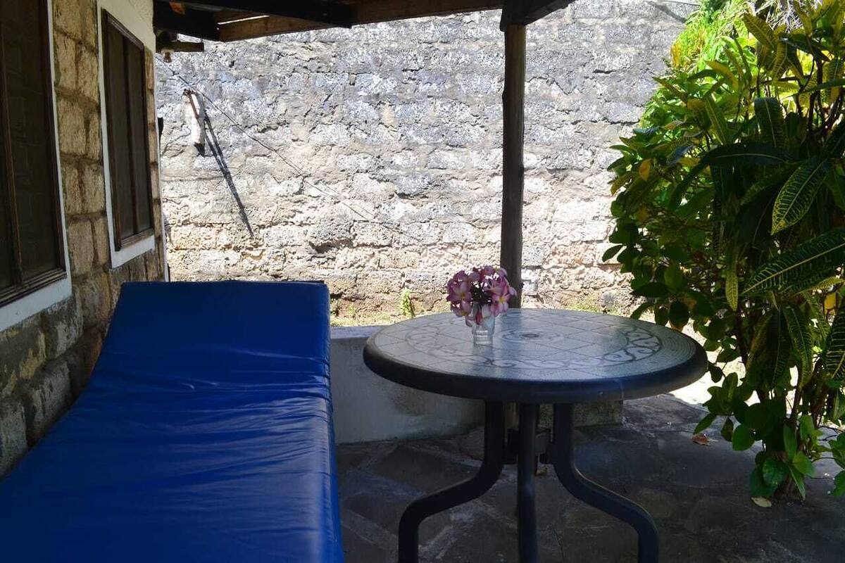 A covered outdoor seating area with a blue cushioned bench, a small round table with a vase of flowers, and a stone wall in the background.
