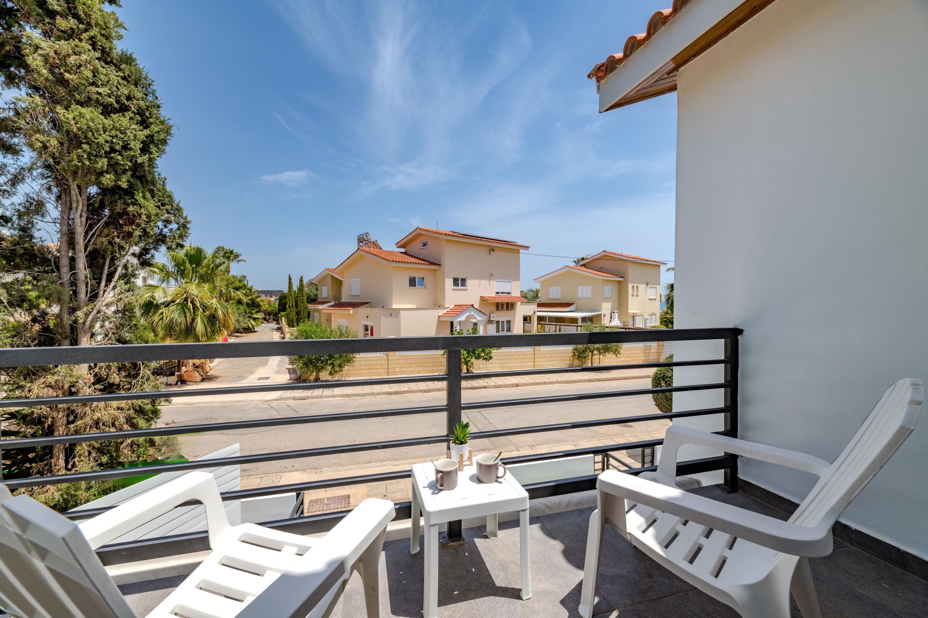 A balcony with white plastic chairs and a small table, overlooking a residential area with houses and a clear sky.