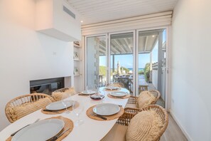 Un coin repas avec une longue table dressée pour six personnes, entourée de chaises en osier, et offrant une vue sur la mer grâce à de grandes portes-fenêtres coulissantes.