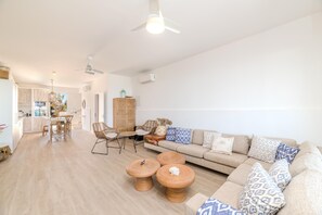 Ein modernes Wohnzimmer mit einer Couch, zwei runden Holztischen, einem Esstisch mit Stühlen und einer Küche im Hintergrund.