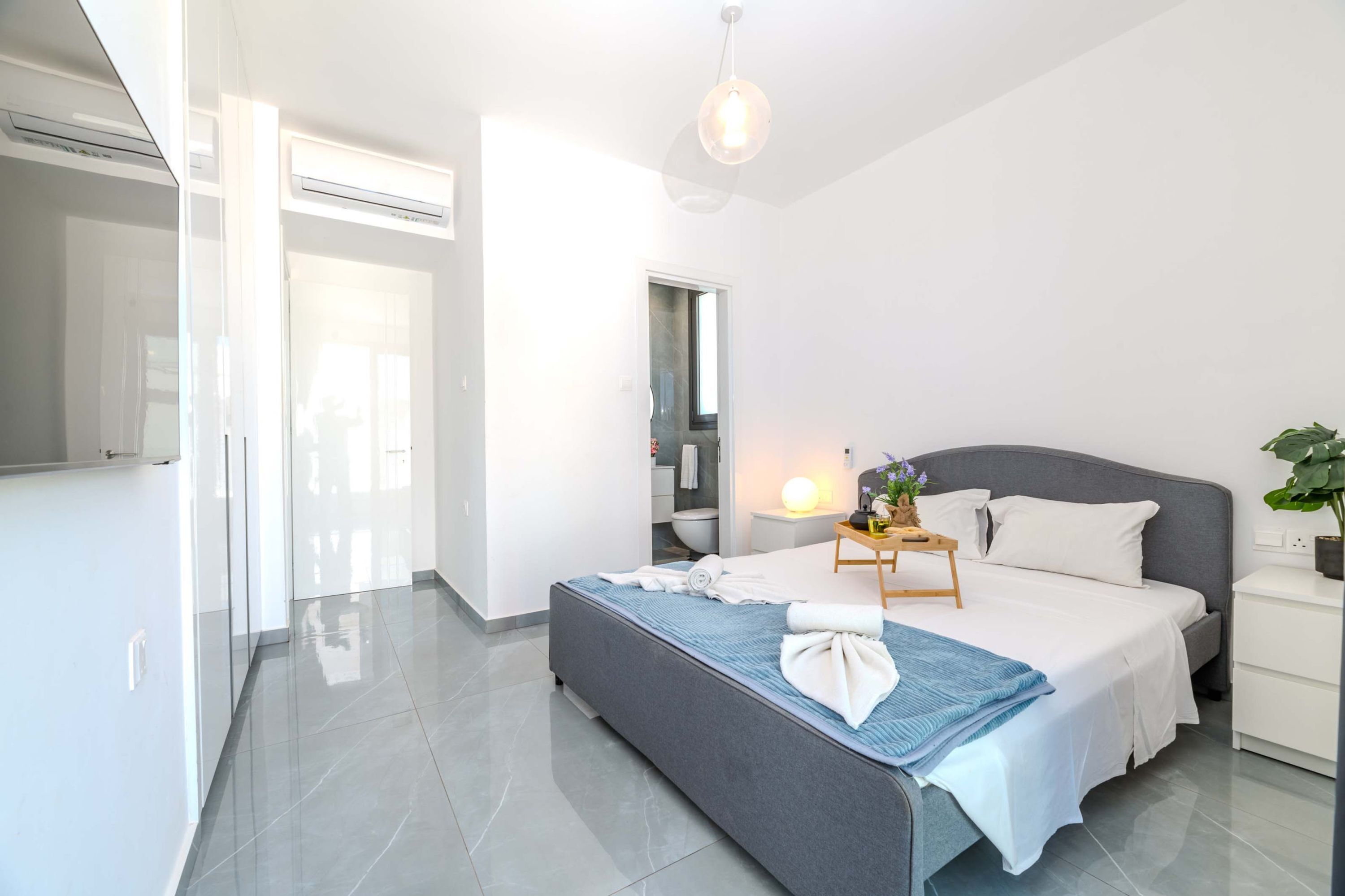 A modern bedroom with a large bed, bedside tables, a nightstand, a mirror, and a hanging light fixture.