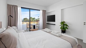 A modern hotel room with a large bed, a flat-screen TV, a small table, and a balcony with a view. - Finikoudes Marina View 202 (Larnaca)