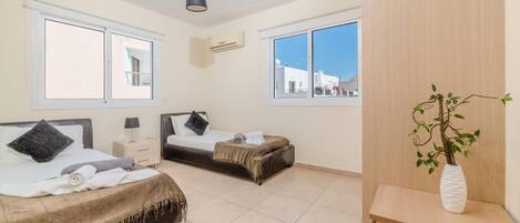 A hotel room with two beds, a bedside table with a lamp, a small plant on a side table, and large windows with a view of buildings.